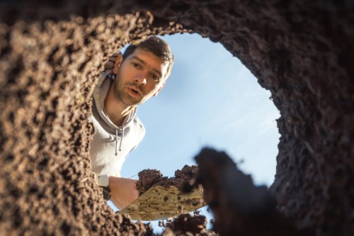 A man looks down into a deep hole, holding a shovel, with blue sky visible above.