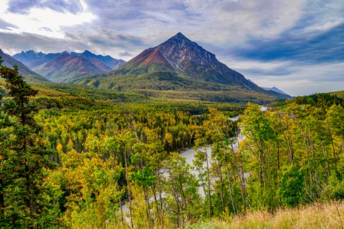 A scenic view of mountains and a winding river surrounded by lush green and autumn-colored trees under a cloudy sky.