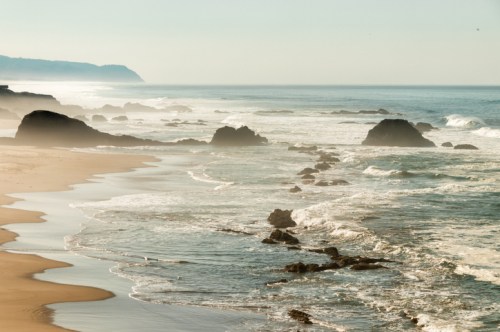 A serene beach scene with gentle waves, rocky formations, and a misty coastline under soft sunlight.