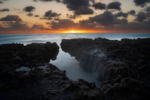 Sunset over the ocean, with rocky formations in the foreground and clouds scattered in the sky.