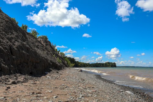 A rocky beach with a steep cliff, under a bright blue sky with fluffy clouds and gentle waves lapping at the shore.
