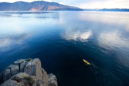 A lone kayaker paddles on calm waters, surrounded by mountains and a serene sky reflecting on the lake's surface.