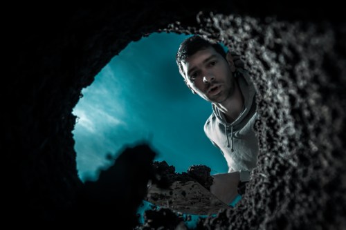 A man looks down into a dark hole, holding a shovel with dirt, illuminated by a blue light.