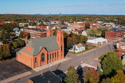 Aerial view of a red brick church with two tall spires, surrounded by trees and buildings in a small town.