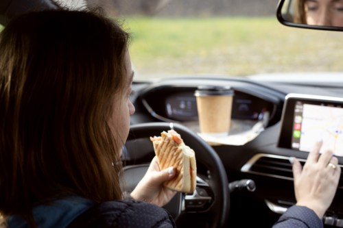 A woman in a car eats a sandwich while using a GPS on the dashboard and a coffee cup is in the cup holder.