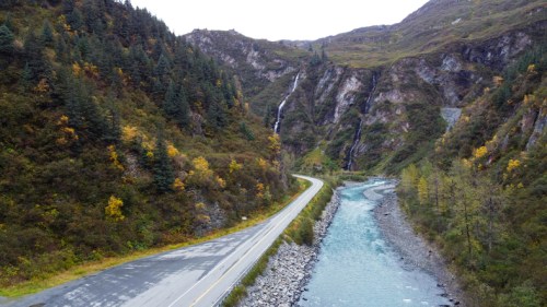 A winding road alongside a river, surrounded by mountains and autumn foliage, with waterfalls cascading in the background.