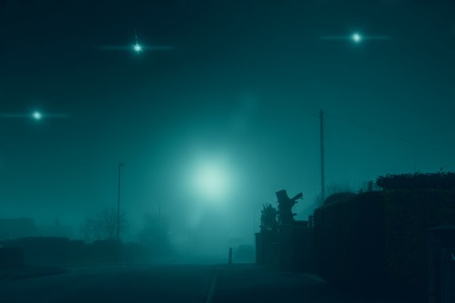 A foggy street scene at night, illuminated by mysterious lights in the sky and a silhouette of a statue.