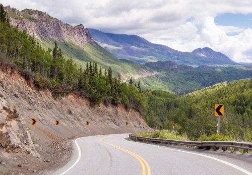 Winding road through lush green mountains under a cloudy sky, with sharp curves and rocky terrain on the side.