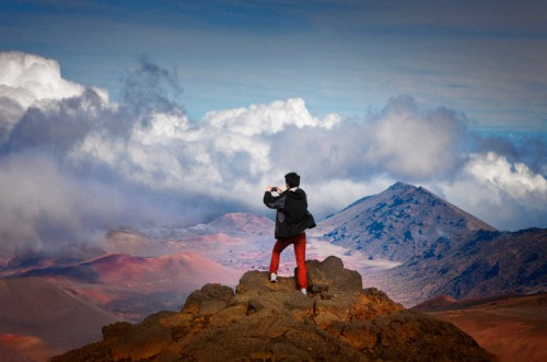 A person stands on a rocky peak, taking photos of a colorful landscape with mountains and clouds in the background.