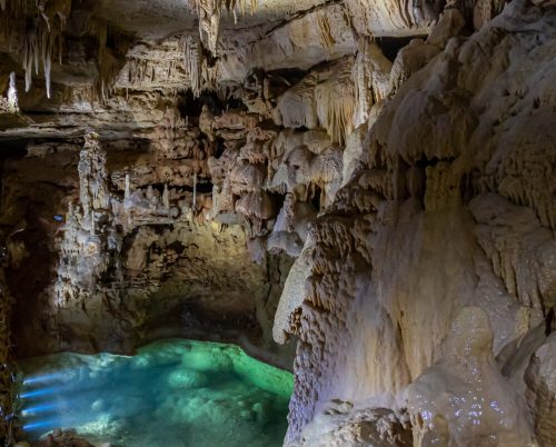 A stunning cave interior featuring stalactites and stalagmites, with a shimmering turquoise pool of water.