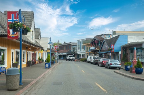 A charming street lined with colorful buildings, shops, and parked cars under a bright blue sky.