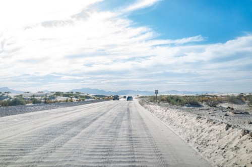 A sandy road stretches into the distance under a bright blue sky with scattered clouds and distant mountains.