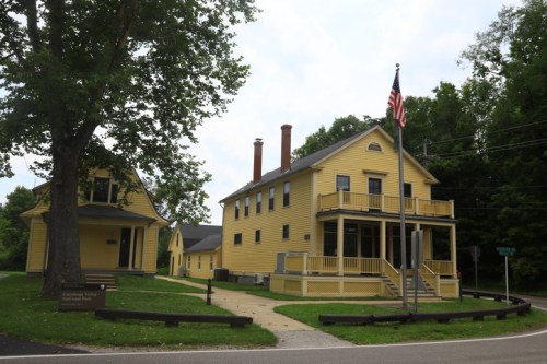 Two yellow houses with a flagpole in front, surrounded by trees and a road. A sign is visible nearby.