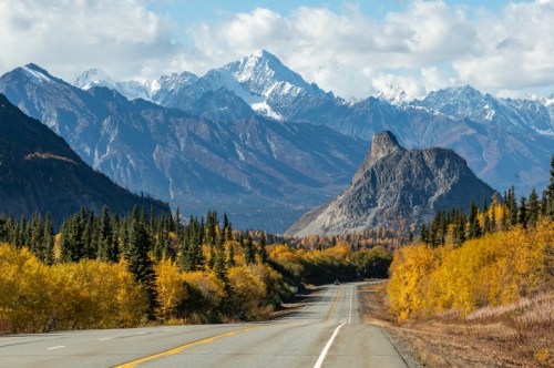 A scenic road winds through vibrant autumn foliage, with snow-capped mountains in the background under a blue sky.
