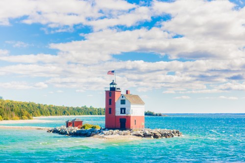 A red lighthouse on a rocky island, surrounded by blue water and green trees under a partly cloudy sky.