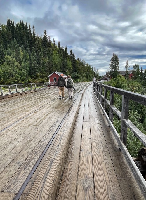 Two people walk along a wooden bridge surrounded by lush greenery and a cloudy sky, with a red cabin in the background.