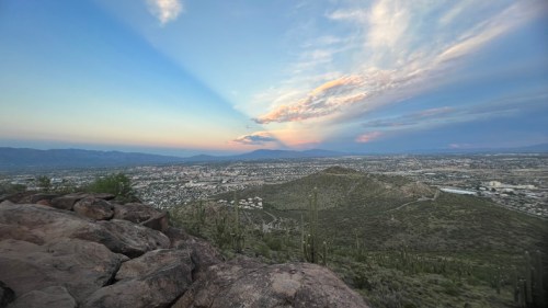 A panoramic view of a desert landscape at sunset, with mountains, city below, and colorful clouds in the sky.