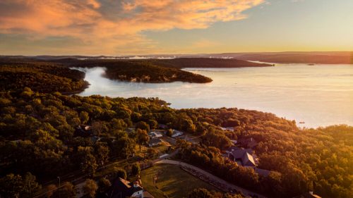 Aerial view of a serene lake surrounded by lush greenery and misty hills at sunset, with homes nestled along the shore.