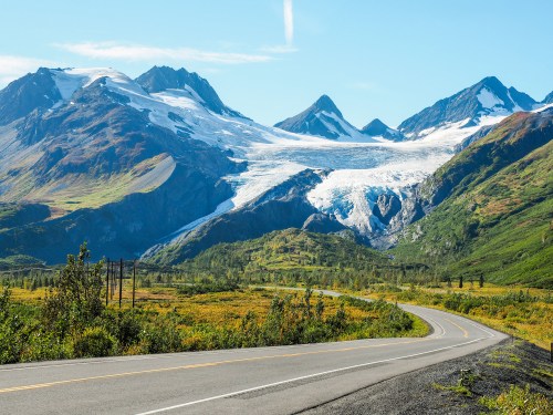 A winding road leads through lush greenery, with majestic mountains and a glacier in the background under a clear blue sky.