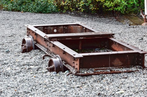 An old, rusted train car frame sits on gravel, surrounded by greenery.