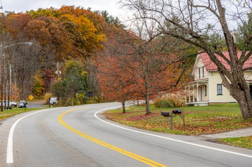 A winding road curves past a house with a red roof, surrounded by autumn foliage and trees.