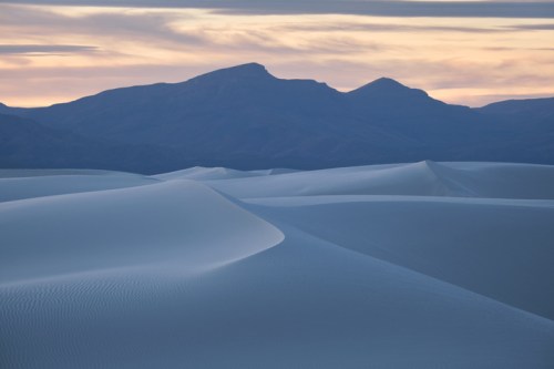 A serene desert landscape with rolling sand dunes and distant mountains under a colorful sunset sky.
