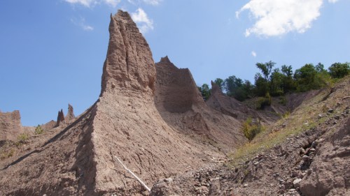 A rugged, towering rock formation rises against a blue sky, surrounded by greenery and steep, eroded slopes.