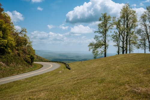 A winding road curves through a grassy hillside with trees, overlooking a distant mountain landscape under a blue sky.