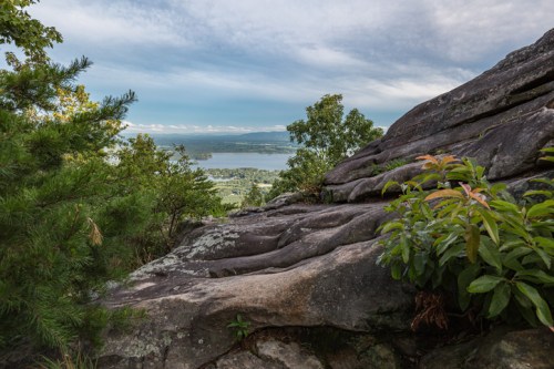 A rocky outcrop with greenery overlooks a serene lake and distant hills under a cloudy sky.