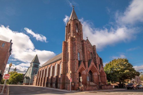 A historic stone church with a tall spire, surrounded by trees and a blue sky with clouds.