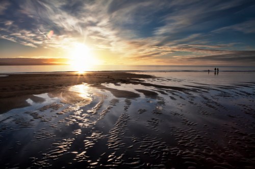 Sunset over a beach with reflective wet sand and silhouettes of people walking along the shore.