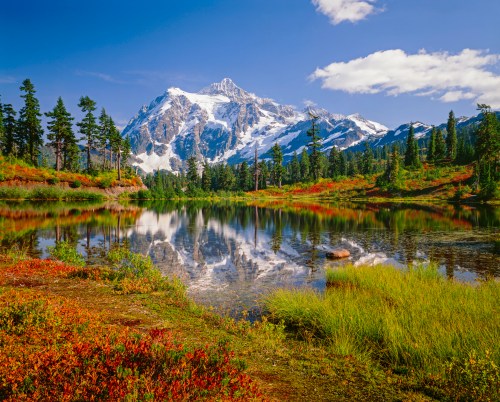A serene mountain landscape with a snow-capped peak reflecting in a calm lake, surrounded by lush greenery and blue skies.