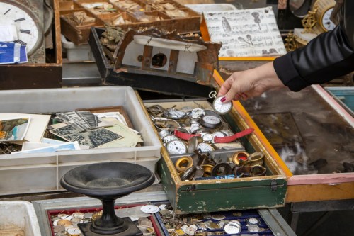 A person examines vintage pocket watches in a cluttered market stall filled with various antiques and collectibles.