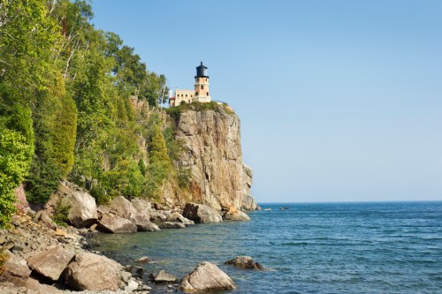 A lighthouse perched on a rocky cliff overlooking a calm blue lake, surrounded by lush green trees under a clear sky.