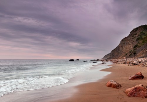 A serene beach scene with gentle waves, rocky shore, and a cloudy sky in the background.