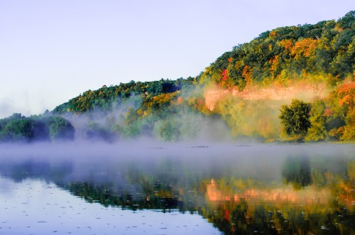A serene river scene with mist, colorful autumn foliage on hills, and reflections in the calm water.