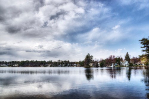 A serene lake scene with reflections of trees and houses under a cloudy sky.