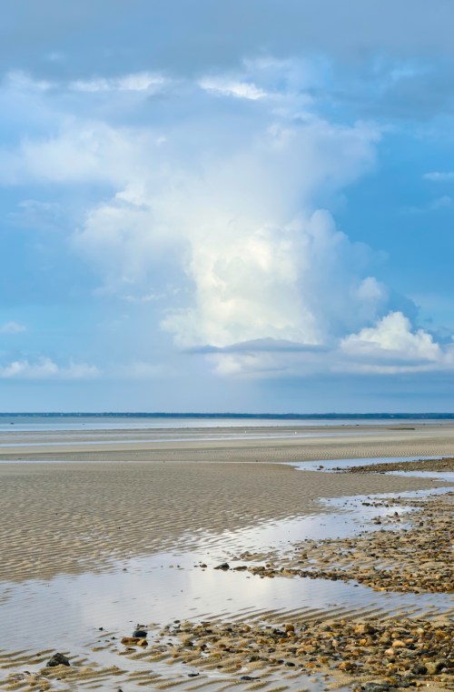 A serene beach scene with wet sand, scattered rocks, and dramatic clouds reflecting in the water under a blue sky.