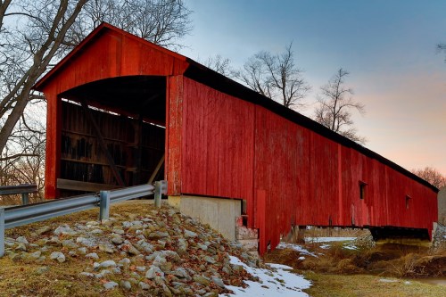 A red covered bridge stands beside a rocky path, surrounded by bare trees and a hint of snow on the ground.