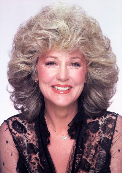 A smiling woman with voluminous, curly hair wearing a black lace top and a butterfly necklace against a light background.