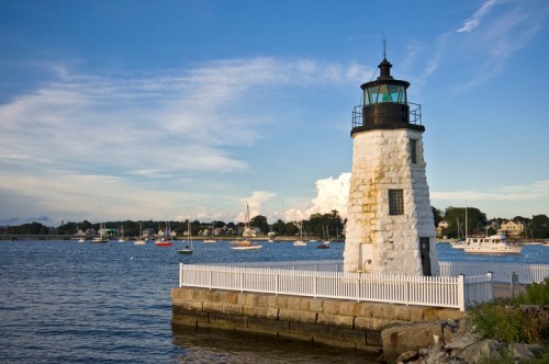 A white lighthouse stands by the water, surrounded by boats and a scenic sky with clouds.
