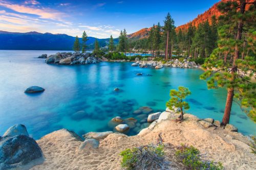A serene lakeside scene with clear blue water, rocky shores, and pine trees under a colorful sunset sky.