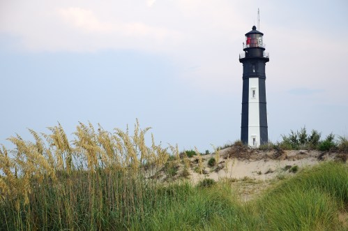 A black and white lighthouse stands on a sandy dune, surrounded by tall grass under a cloudy sky.