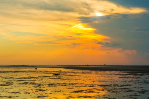 A serene beach at sunset, with vibrant orange and yellow hues reflecting on the wet sand and distant silhouettes of people.