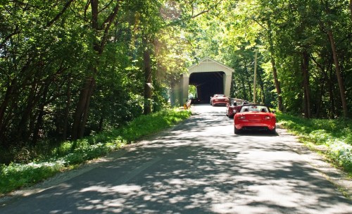 A scenic road leads to a covered bridge, with several red cars parked along the tree-lined path.