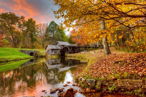 A wooden mill by a tranquil pond, surrounded by vibrant autumn foliage and a colorful sunset sky.