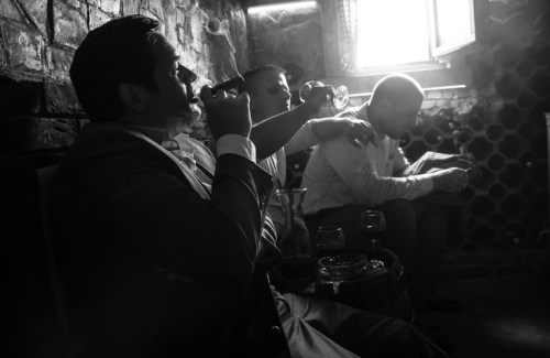 Three men in a dimly lit room, enjoying drinks and cigars, with a vintage ambiance and brick walls.