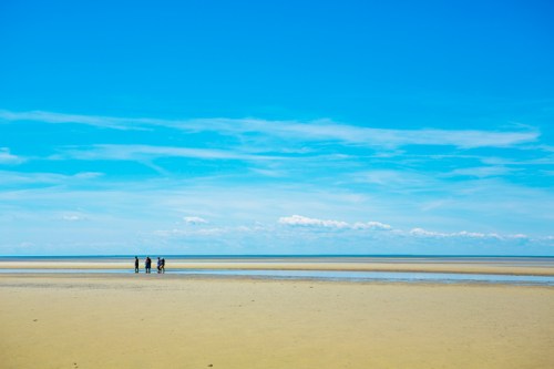 Four people walking on a sandy beach under a bright blue sky with scattered clouds.