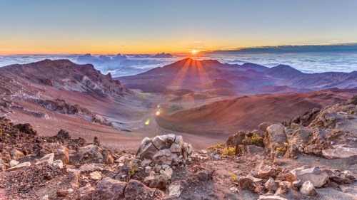 A stunning sunrise over a mountainous landscape, with rocky terrain and clouds below, creating a serene atmosphere.