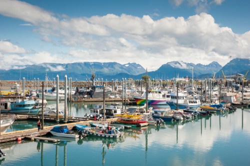 A bustling marina filled with various boats, surrounded by mountains and a clear blue sky with scattered clouds.